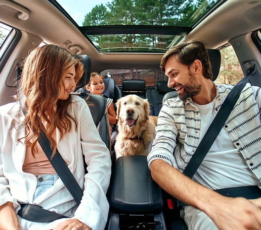 Happy family with a child and a dog traveling together in a car, parents in the front seats smiling.