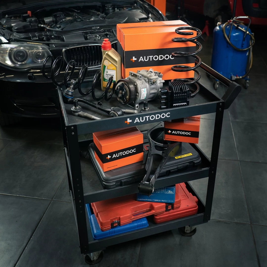 Autodoc car parts on a workshop trolley, including shock springs, compressor, oil, and tools in front of a car.