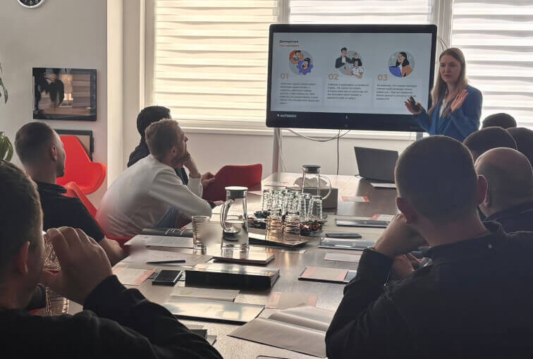 A woman standing in front of a large table with people sitting around it
